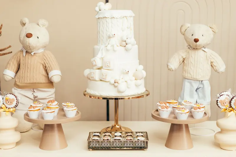 A white tiered cake with cupcakes and two teddy bears on a baby shower table