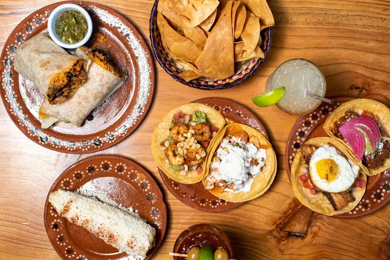Tacos, a burrito, chips, salsa and a cold drink spread across a wooden table