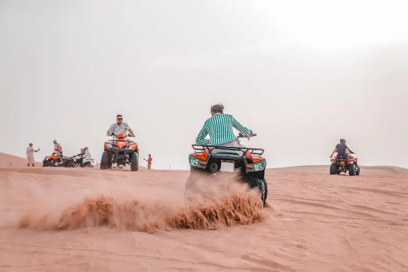 Personas en cuatrimotos levantando arena sobre una duna con sol fuerte