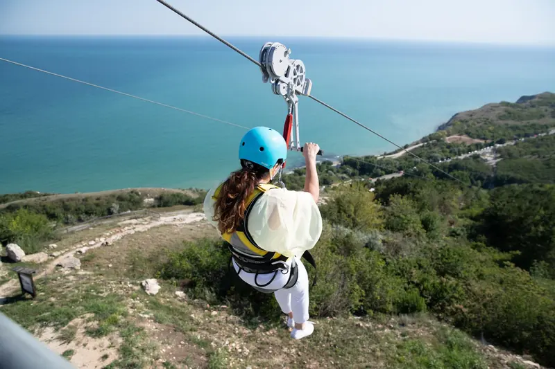 A rider in a blue helmet ziplining above green hills with the sea behind