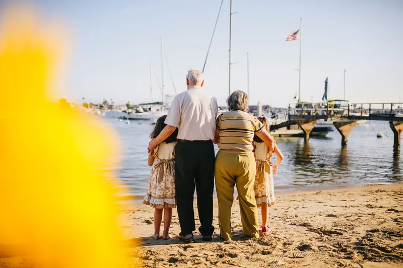 Grandparents and two children standing together on a beach by a dock with boats behind