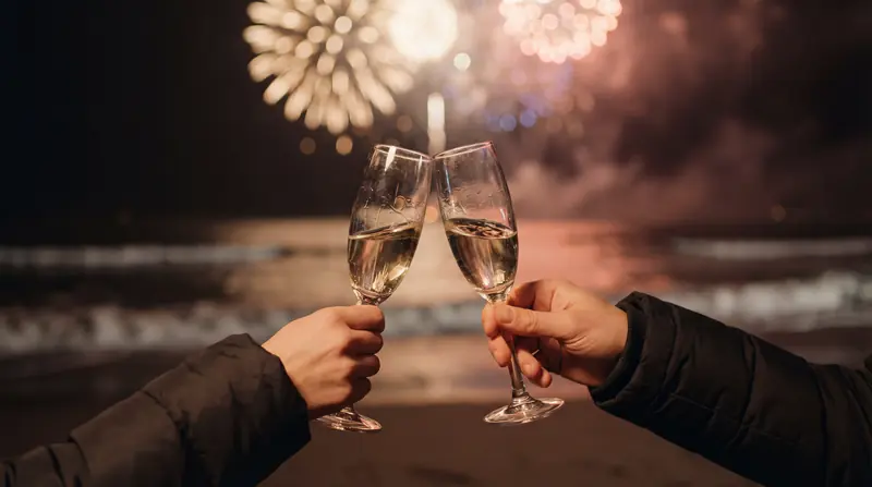 Dos personas brindando con copas de champaña en una playa con fuegos artificiales al fondo