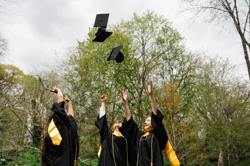 Three graduates in gowns throwing their caps into the air under trees