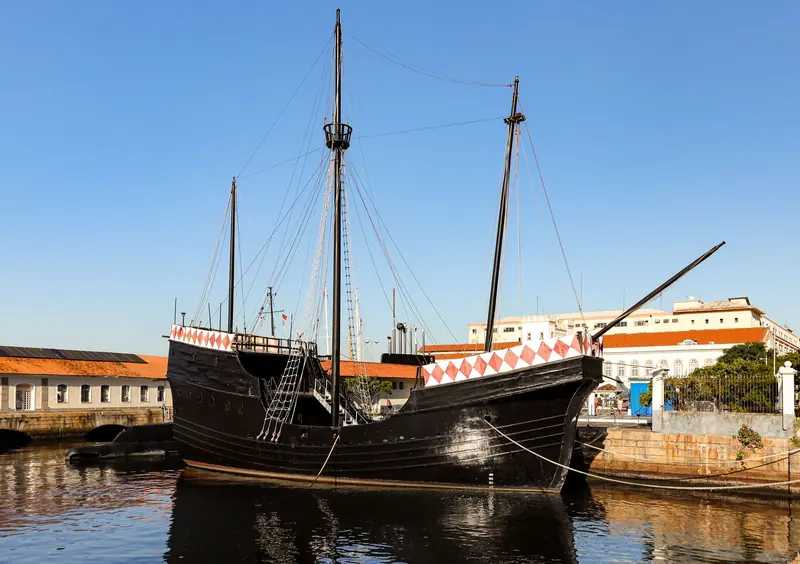 Un velero de madera negro con mástiles altos amarrado en un puerto tranquilo, hoy una réplica de época