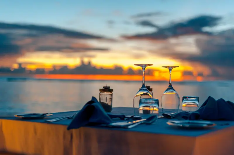 A dinner table set with wine glasses and napkins by the sea as the sun goes down