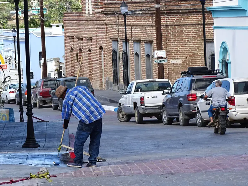 Downtown Todos Santos street with brick buildings, parked cars and a man sweeping the sidewalk
