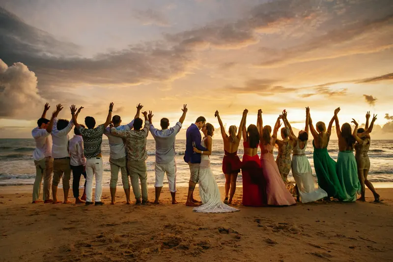 Novios y su grupo de invitados de espaldas en la playa, con los brazos arriba al atardecer