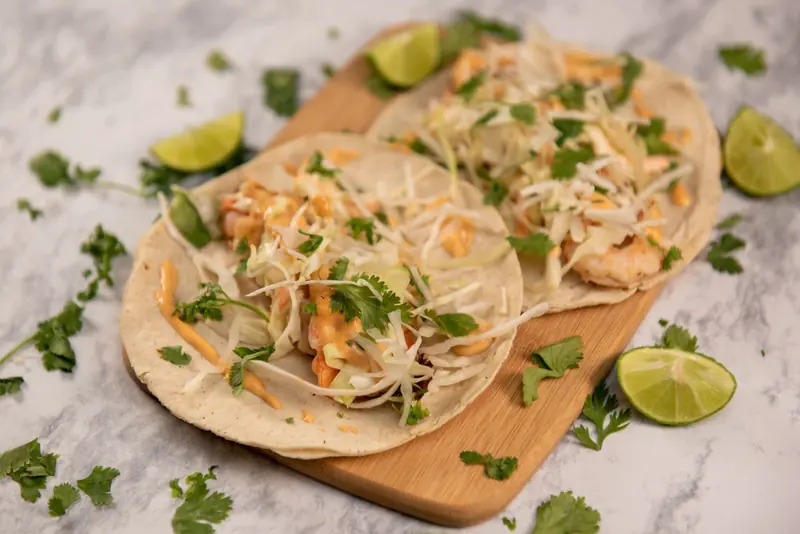 Tacos de camarón con col rallada y cilantro sobre una tabla de madera, con limones partidos