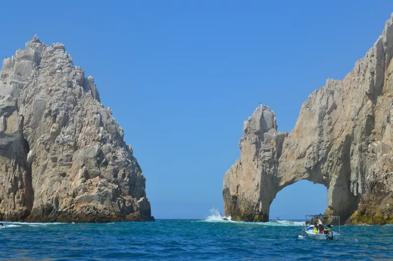 A small boat passing beneath the granite Arch at Land's End in Cabo San Lucas, with a tall rock pinnacle to the left