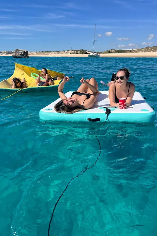 Dos invitadas recostadas en la plataforma flotante del Sea Ray 55, con amigas en el tapete y la playa atrás